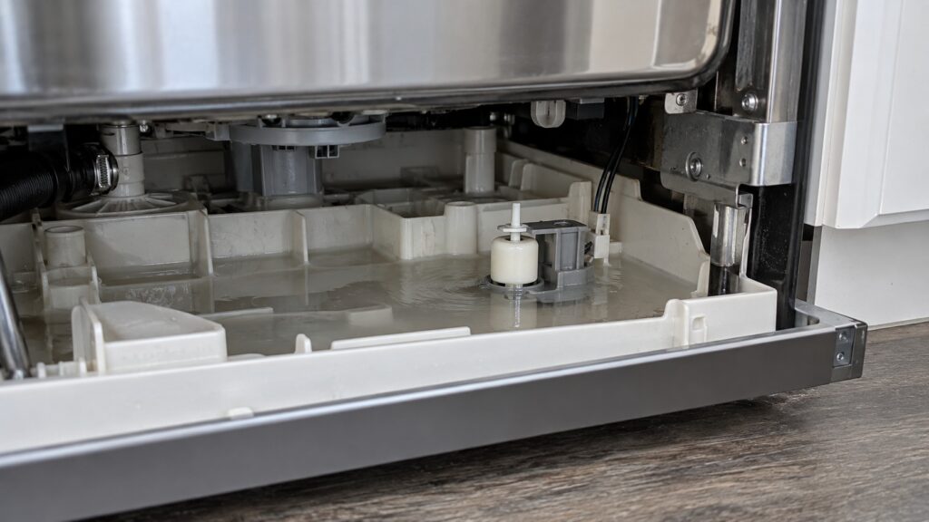 The molded plastic base tray under a dishwasher drawer with a small float switch sitting in shallow water