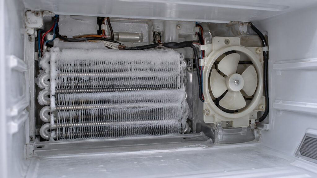 The rear wall of a freezer compartment with the panel removed, showing a frosted evaporator coil and the circulation fan beside it